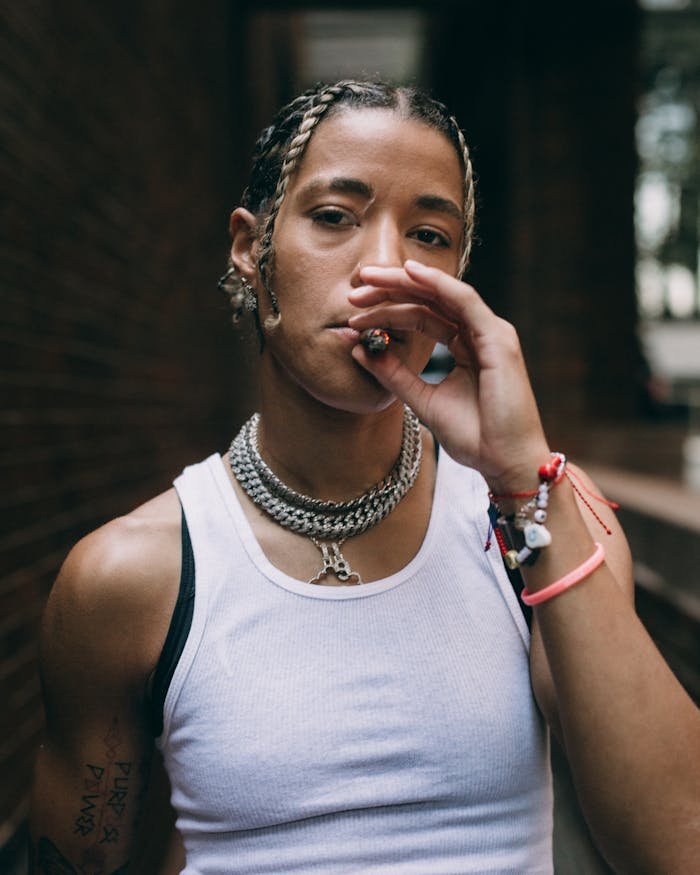 Portrait of a young woman with braided hair smoking outdoors, wearing a white tank top and chains.